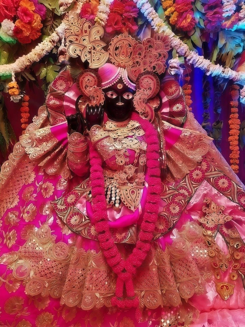 Dark-stone deity idol in magenta and gold embroidered lehenga with golden headdress, pink garland and raised blessing hand
