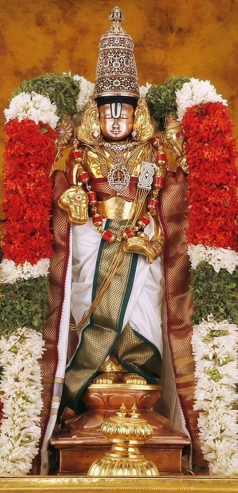 Lord Venkateswara idol in gold armour and tall crown, draped in red and white garlands, on a lotus base